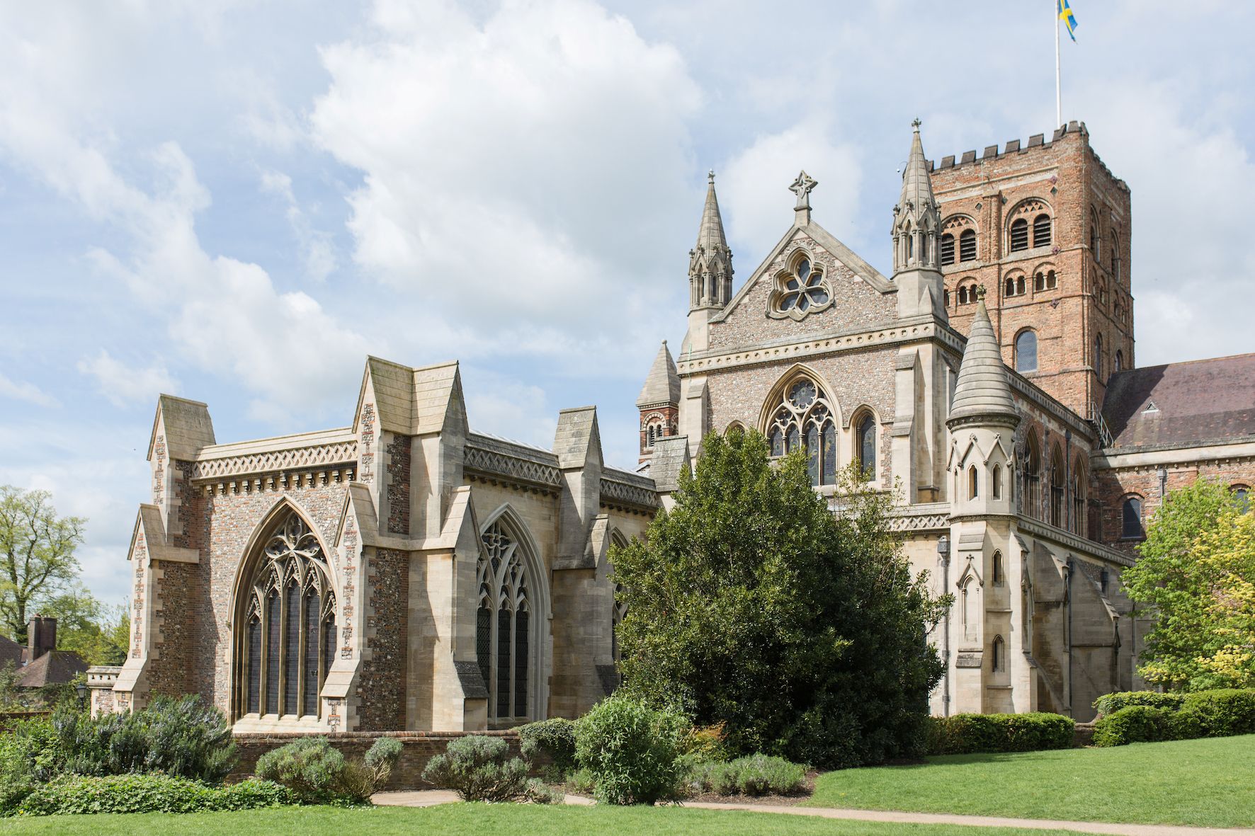 St Albans Cathedral rising above Vintry Gardens, with clipped lawns and greenery in the foreground beneath a bright, cloud-filled sky.
